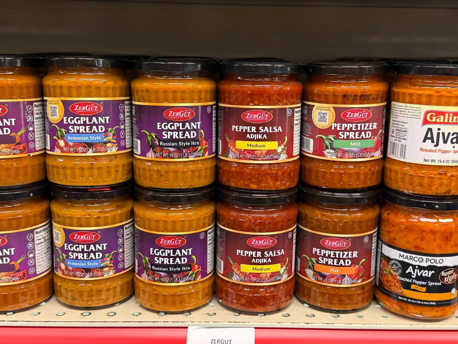 Jars of ajvar, eggplant spread, and pepper salsa lined up on the shelf